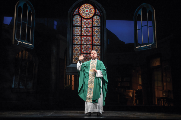 The image is a picture of a priest on a stage with a green robe, with a stained glass window in the back
